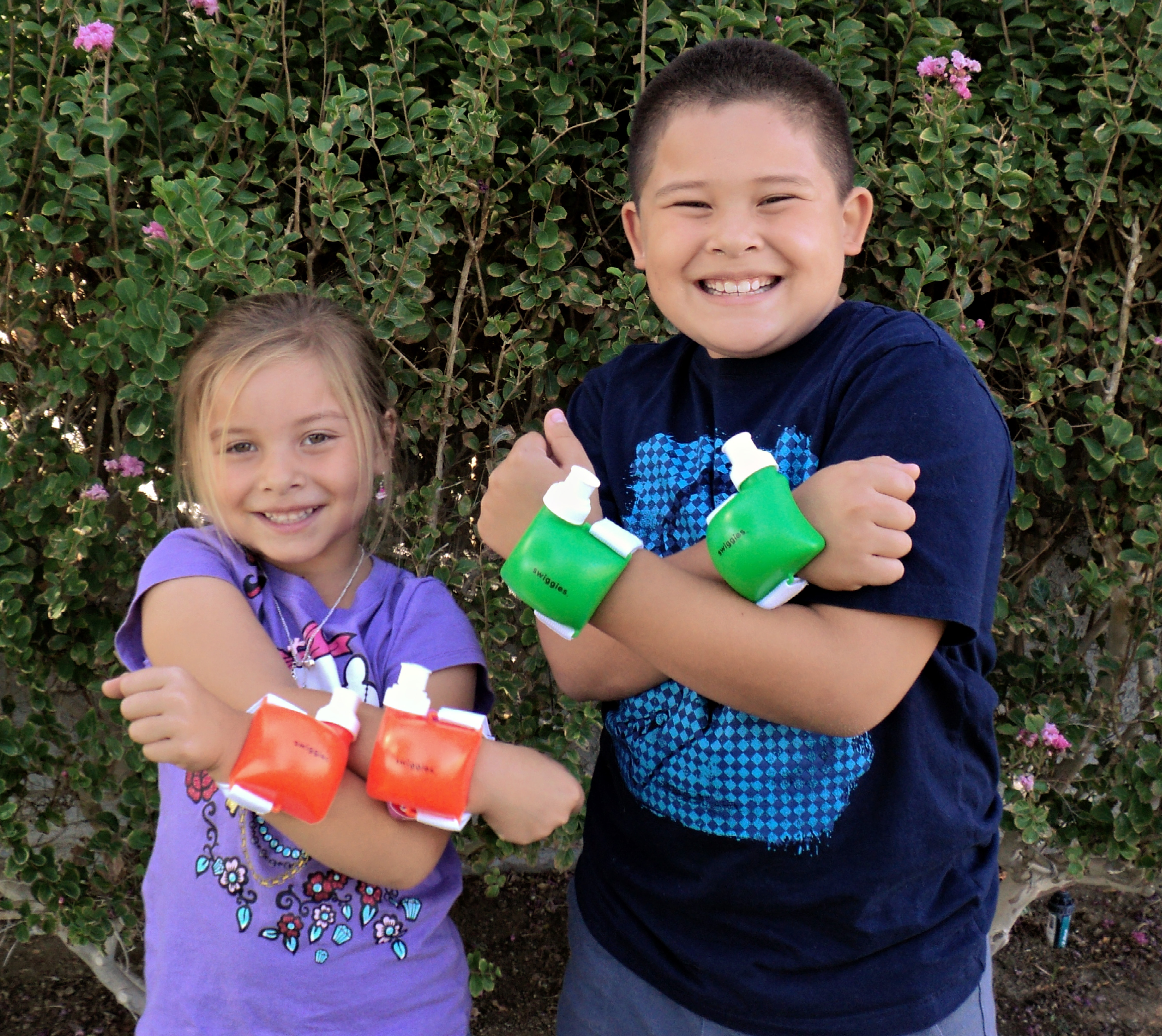 Two smiling kids outdoors, each wearing a pair of Swiggies wrist water bottles — one set in green, the other in red-orange.